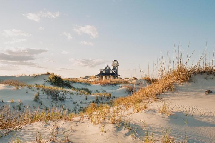A house with a tower stands among sand dunes covered in dry grass under a partly cloudy sky.