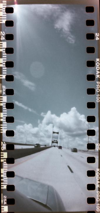 A bridge is visible in the distance from a car driving on a highway under a cloudy sky.