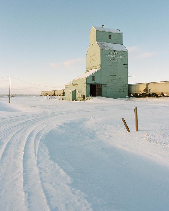 A light green grain elevator stands in a snow-covered field with train cars behind it.