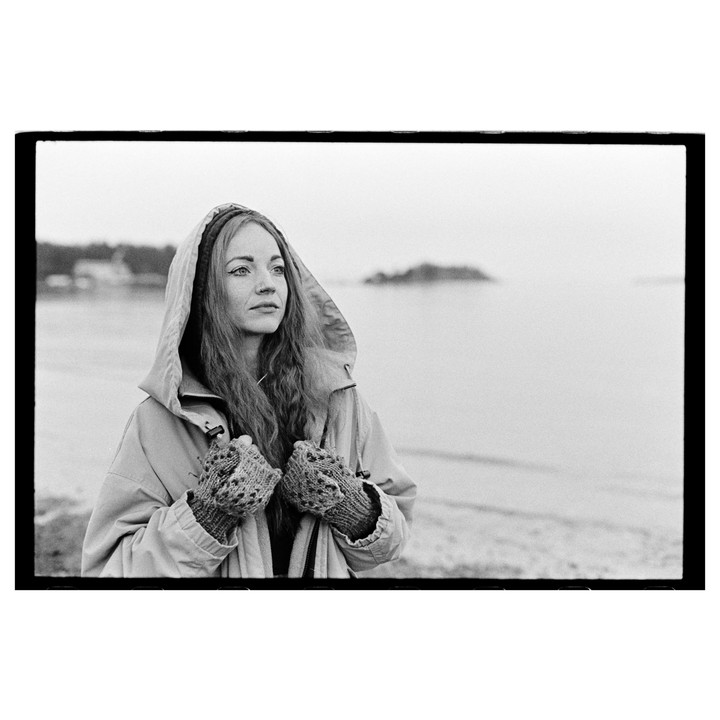 A woman wearing a hooded coat and mittens stands on a beach looking out at the water.