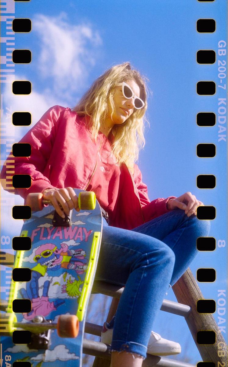 A woman in a red jacket and sunglasses sits with a skateboard against a bright blue sky.