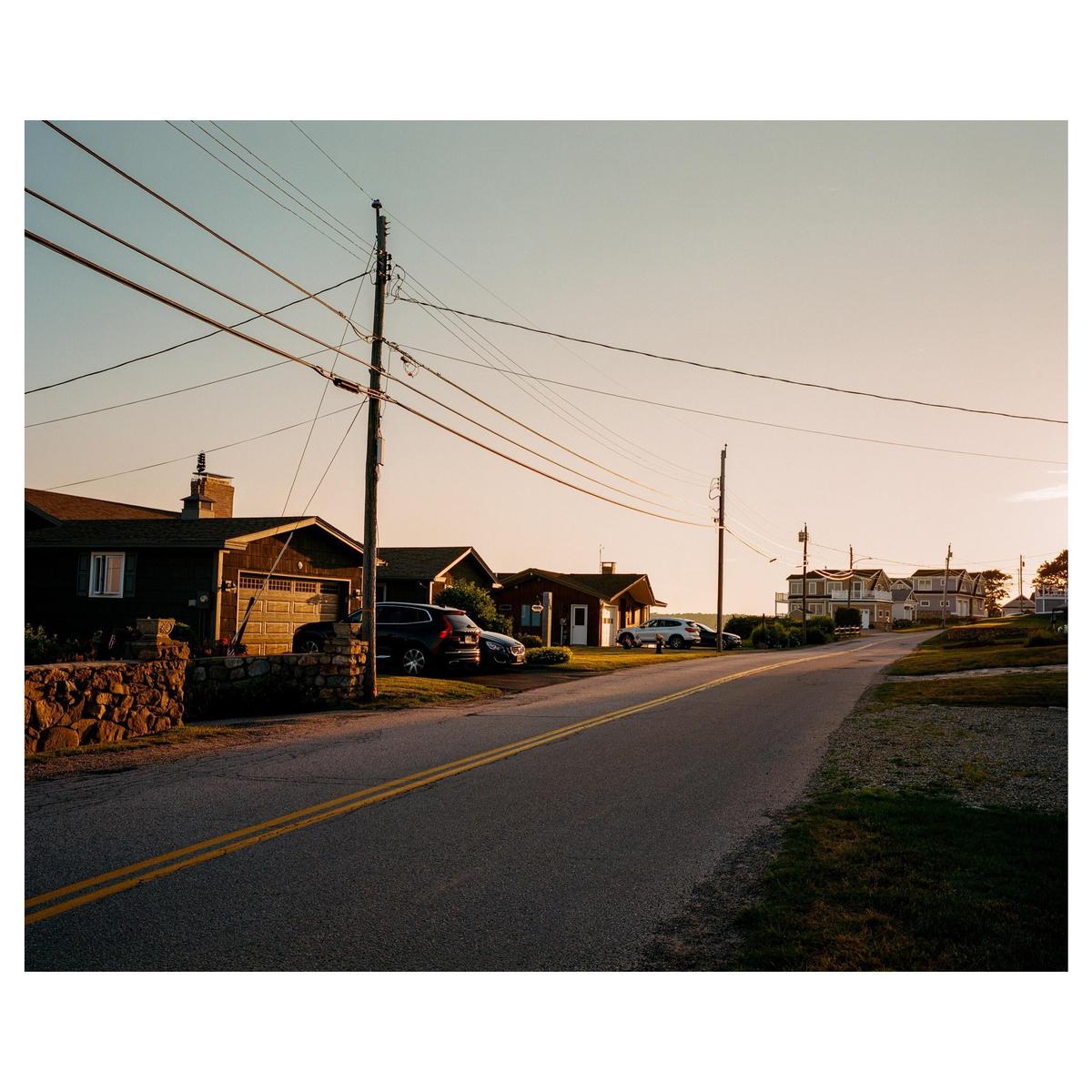 A road with double yellow lines curves past houses and power lines under a golden hour sky.