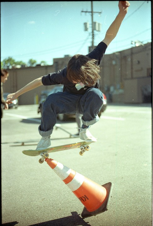 A skateboarder jumps over an orange traffic cone in a parking lot.
