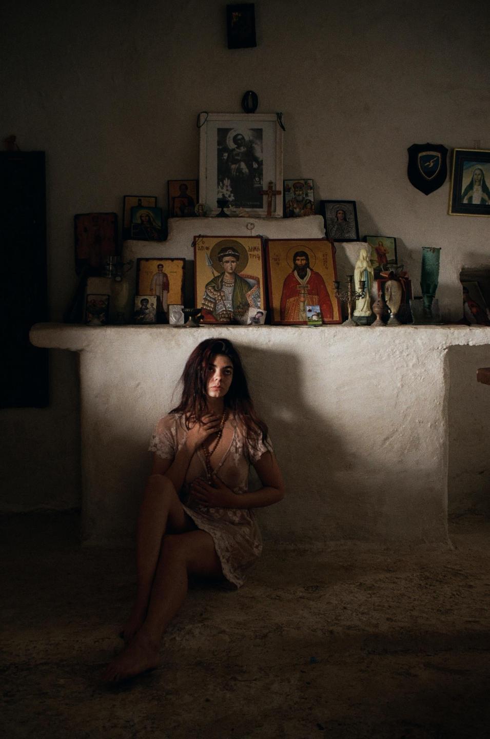 A woman sits on the floor in a room with religious icons on a shelf behind her.