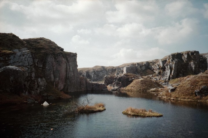 A body of water is surrounded by rocky cliffs and grassy hills under a cloudy sky.