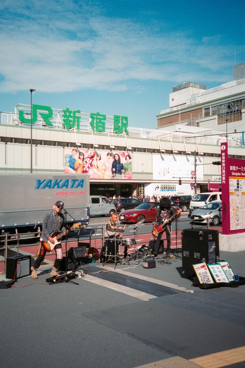 A band performs on a street in front of a train station with cars and trucks passing by.
