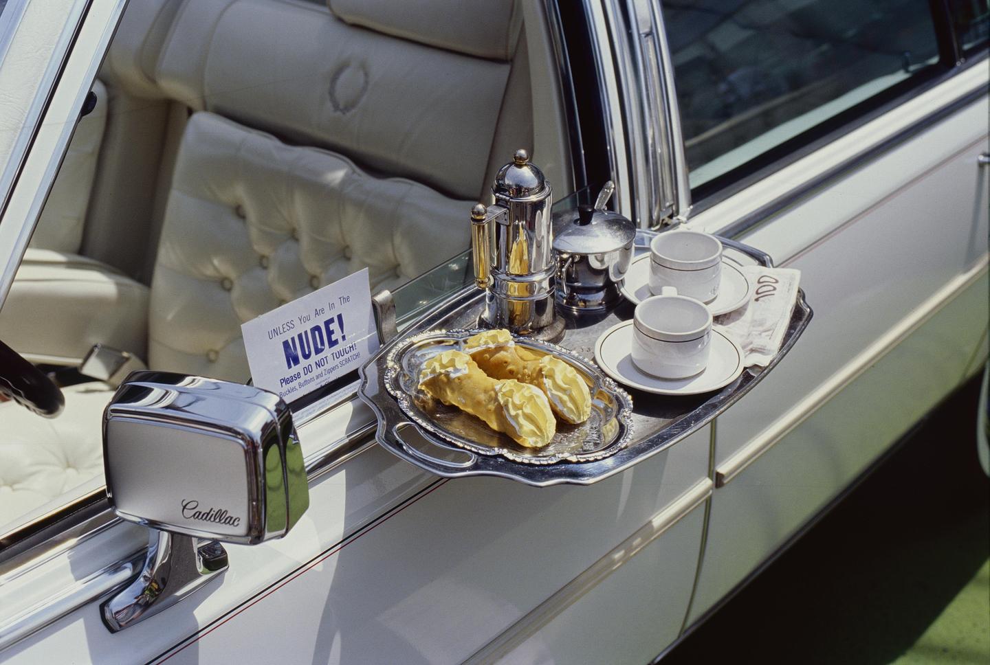 A silver tray with cannoli, coffee cups, and a coffee pot is attached to the side of a white Cadillac.