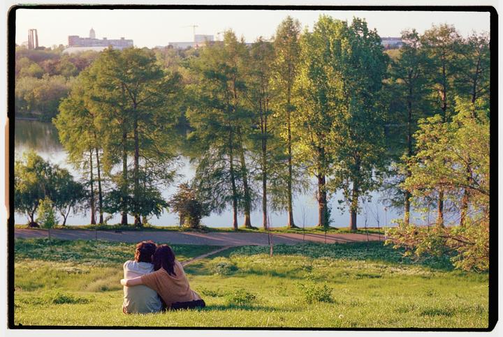A couple sits on a grassy hill overlooking a lake lined with trees and distant buildings.