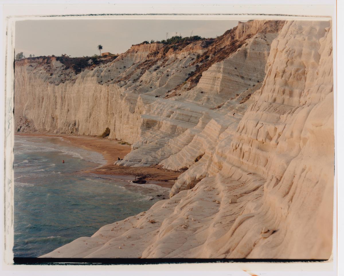 A sandy beach is nestled at the base of towering white cliffs, with a single palm tree visible at the top of the cliff.