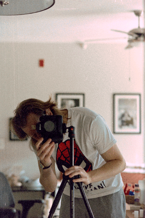 A person with long hair wearing a white t-shirt with a Spider-Man logo is taking a self-portrait in a mirror with a camera on a tripod.