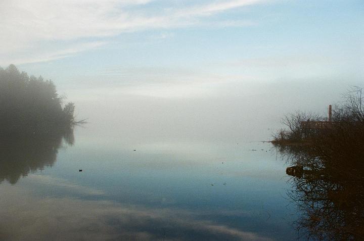 A body of water reflects the sky and trees on a hazy day, with a building and chimney visible on the right bank.