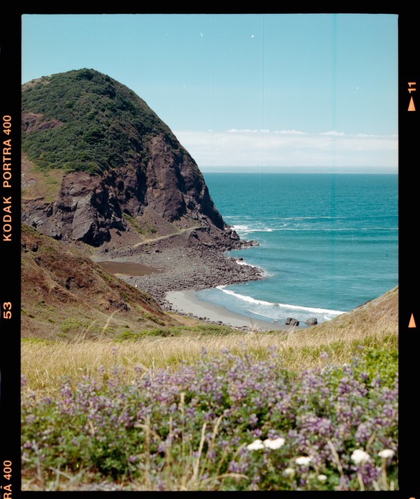 A rocky mountain covered in green foliage overlooks a sandy beach and the ocean under a clear sky.