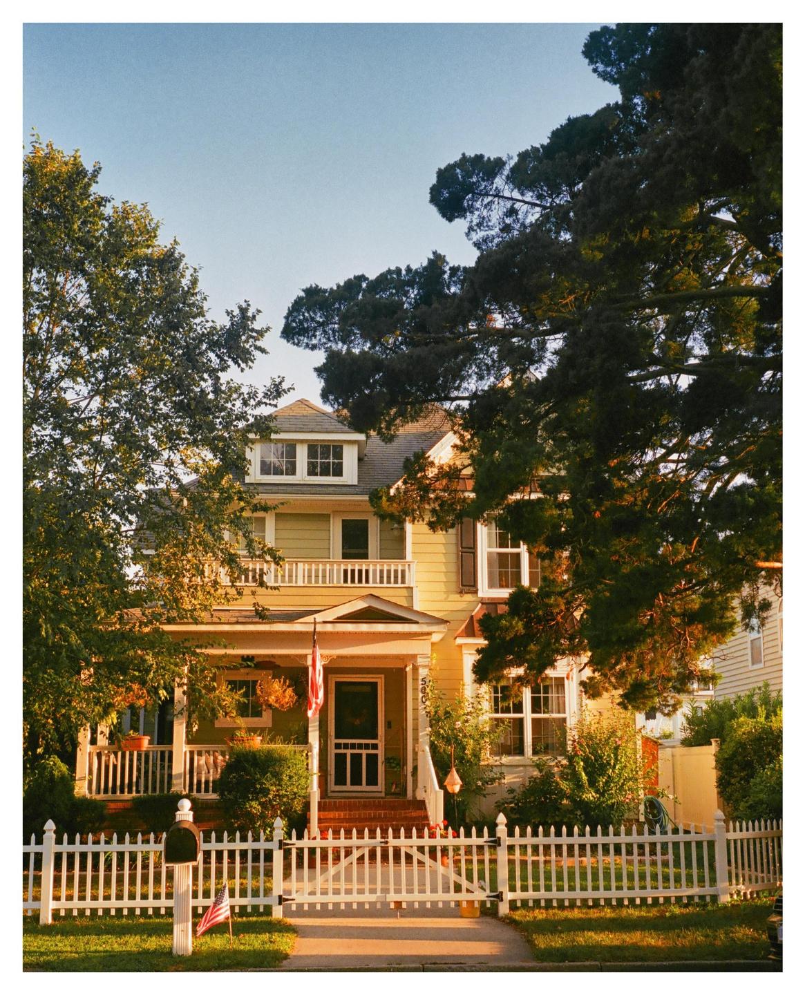 A yellow house with a white picket fence and an American flag hanging from the porch is surrounded by trees.
