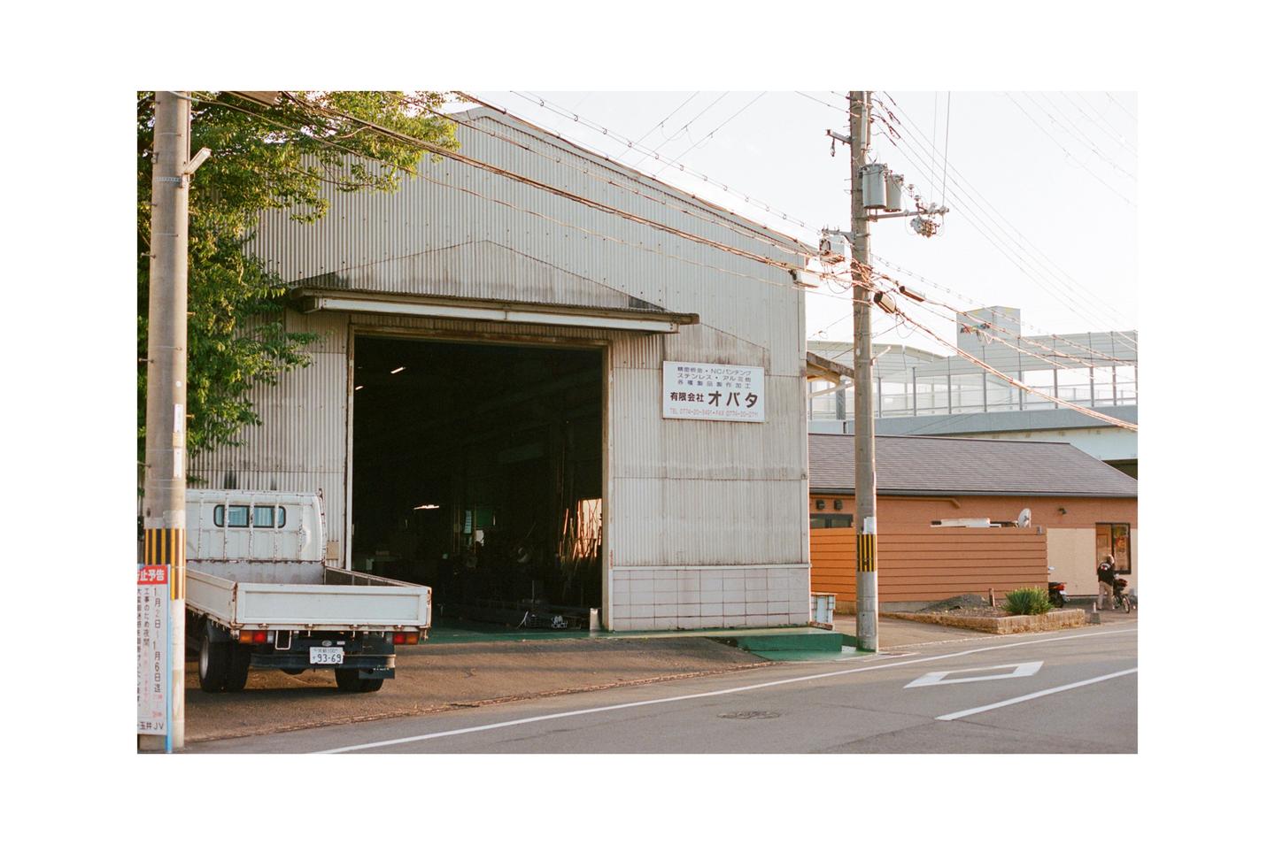 A white flatbed truck is parked in front of a large industrial building with its garage door open, next to a street with power lines overhead.