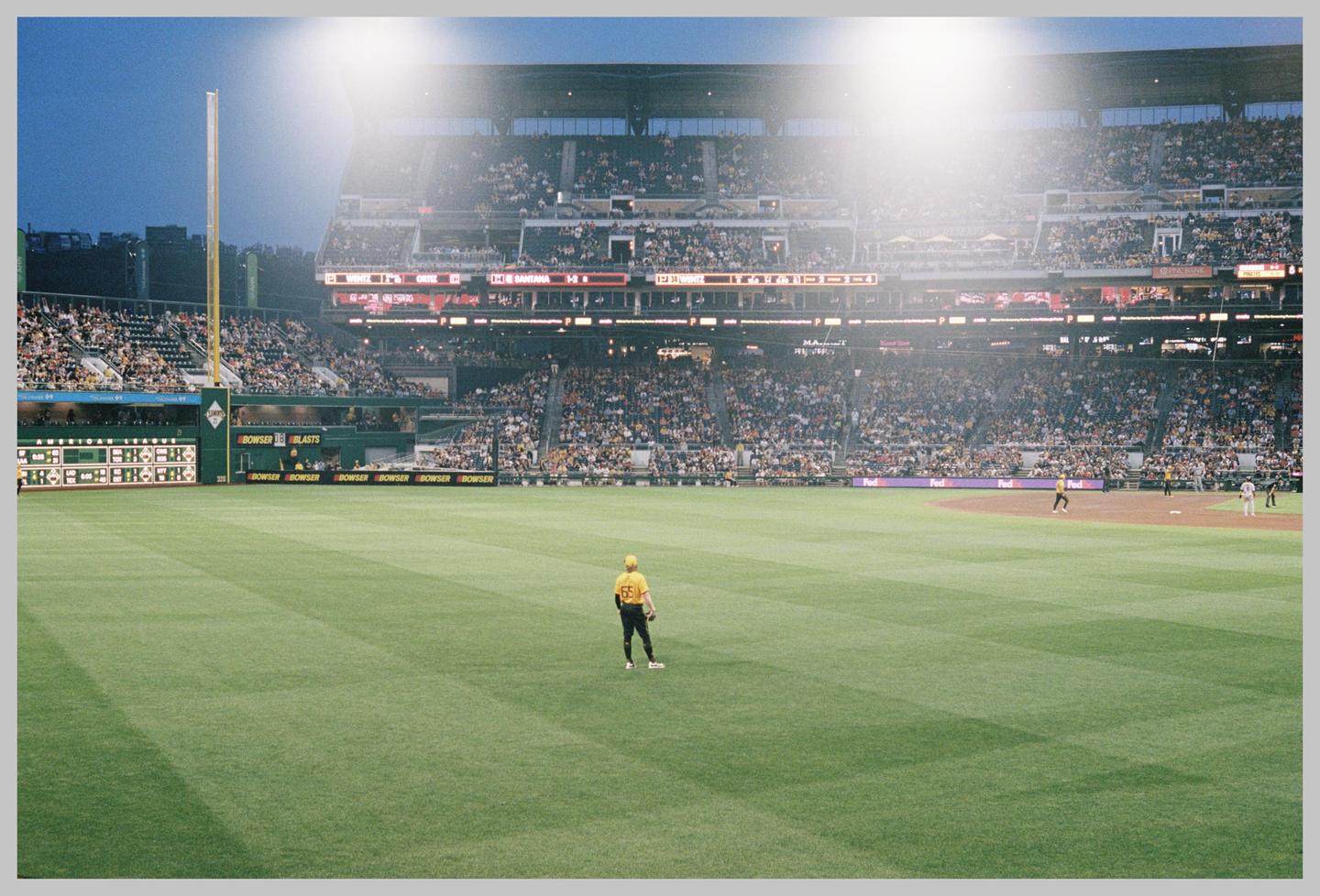 A baseball player stands on a green field in a stadium filled with spectators under bright lights.