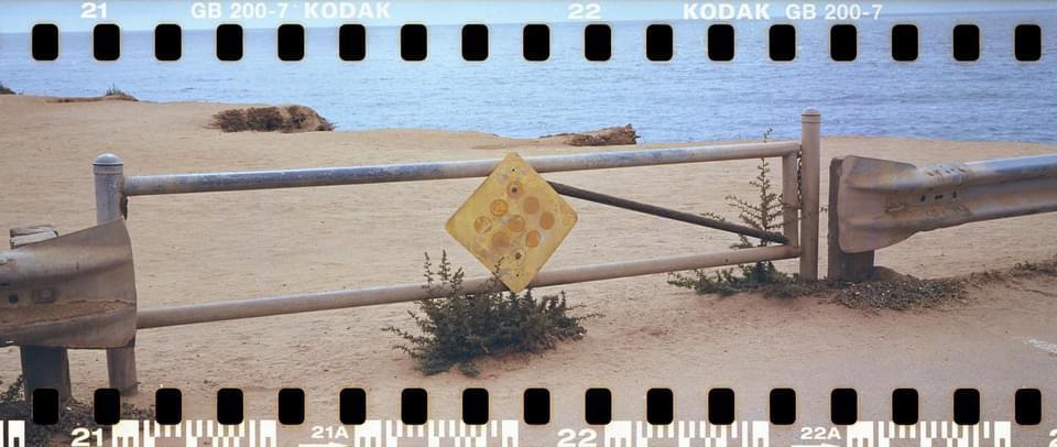 A yellow diamond-shaped sign with circular holes is attached to a metal fence on a sandy beach next to the ocean.