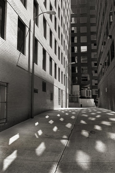 Sunlight casts square patterns on the ground of a narrow alleyway between two tall buildings.