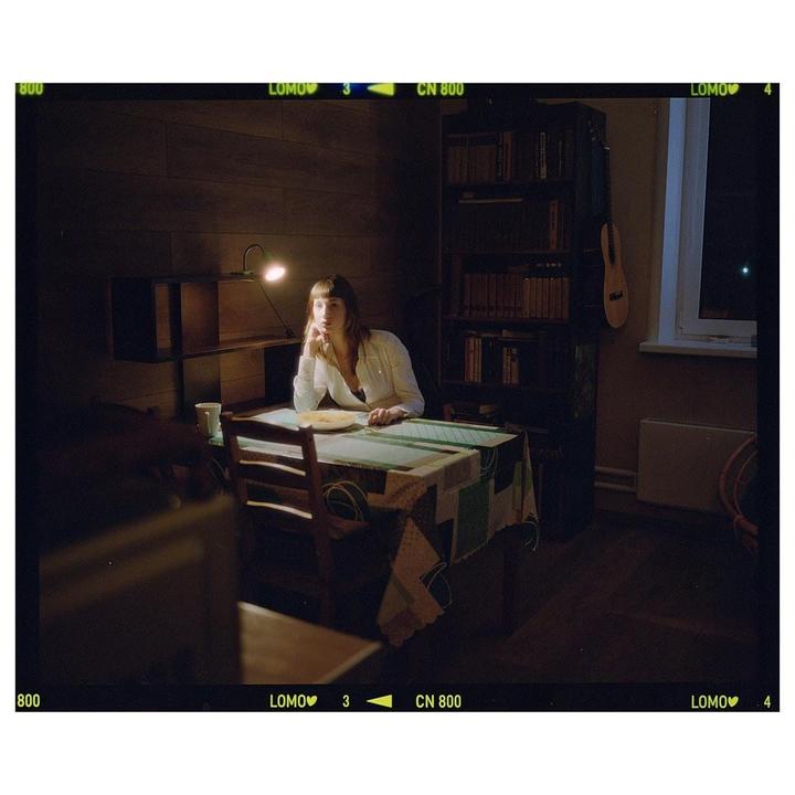 A woman sits at a table with a plate of food, illuminated by a desk lamp, in a dimly lit room.
