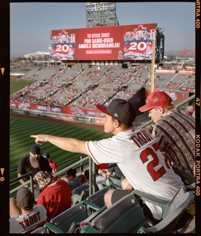 A man in a baseball jersey points towards the field from the stands of a baseball stadium.