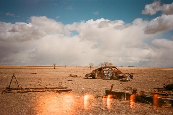A rusted, abandoned car with "URON SACRED LAND" painted on its side sits in a dry, open field under a cloudy sky.