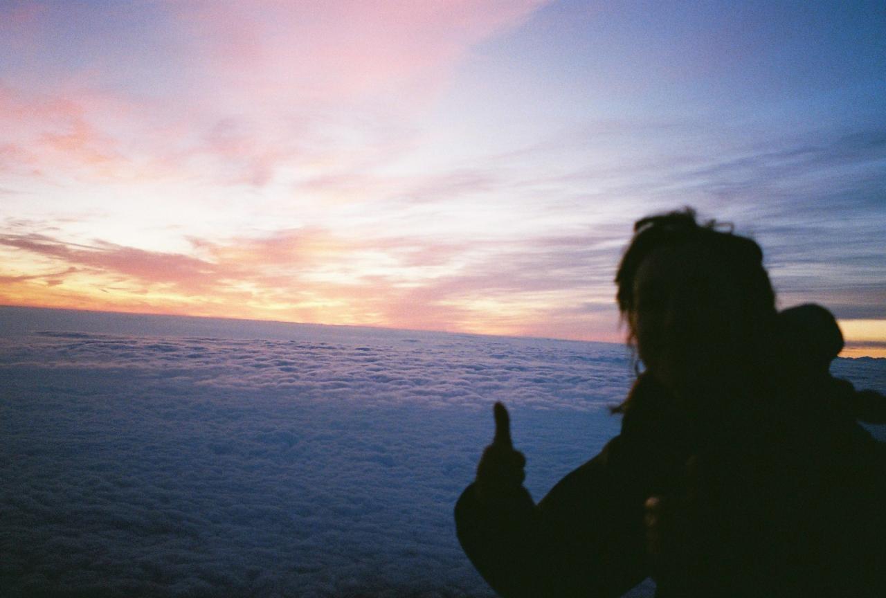 A person is silhouetted against a colorful sky above a sea of clouds, giving a thumbs up.