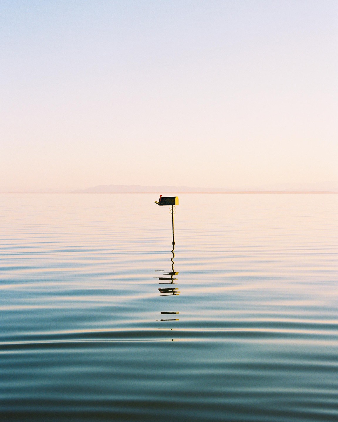 A mailbox stands in the middle of a calm body of water under a clear sky.