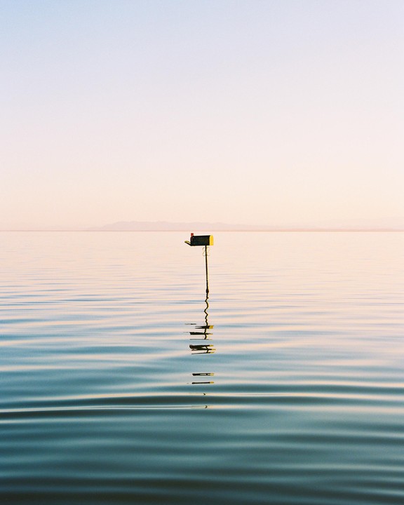 A mailbox stands in the middle of a calm body of water under a clear sky.