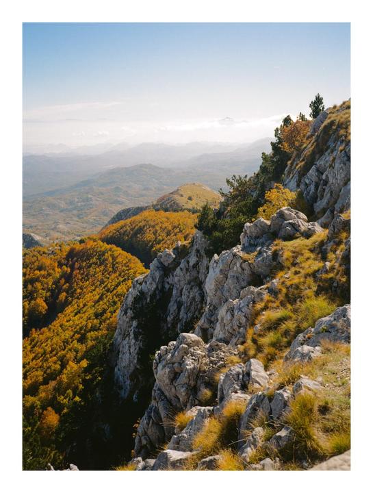A mountain landscape shows rocky slopes and valleys filled with autumn trees under a hazy sky.
