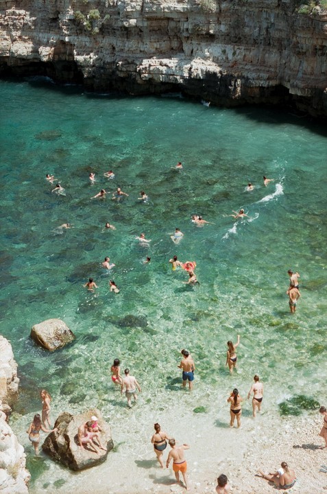 A high-angle view shows many people swimming and standing in clear turquoise water next to a rocky cliff.