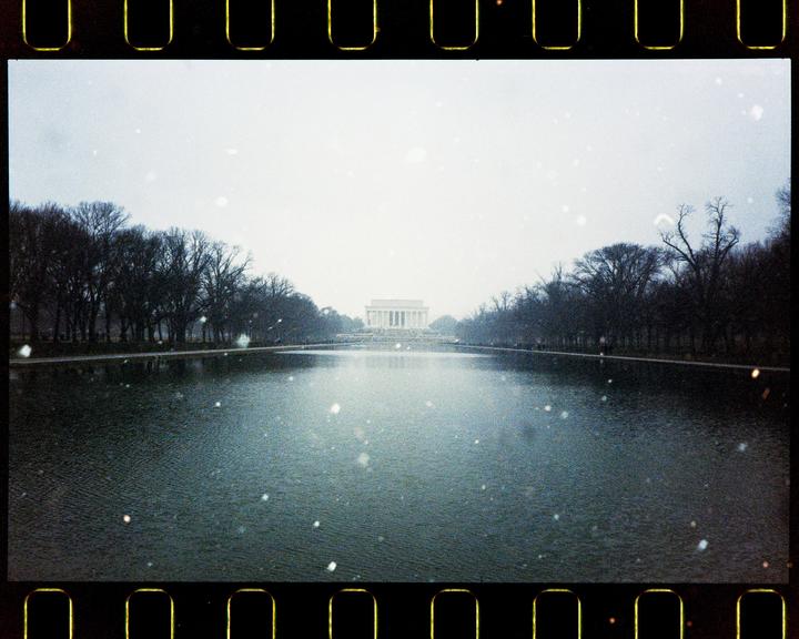 The Lincoln Memorial is visible in the distance across a body of water, with trees lining both sides of the water under a snowy sky.