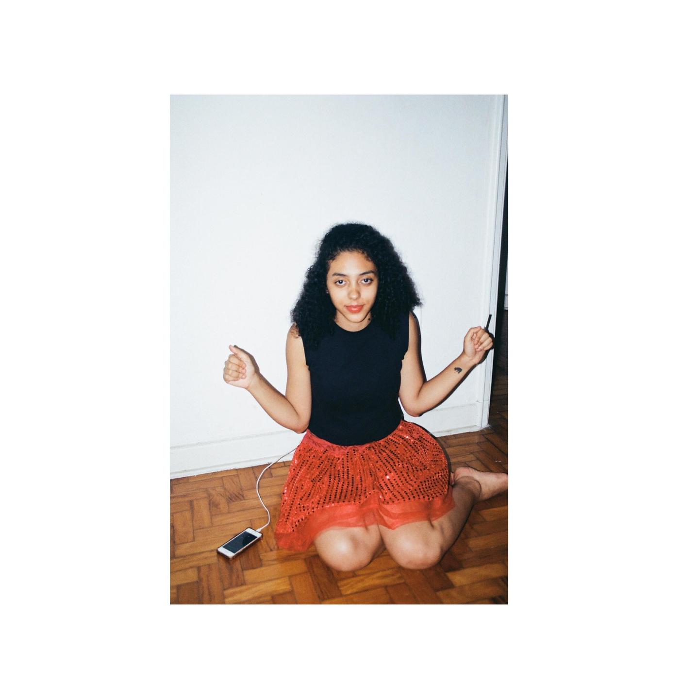 A woman with curly hair kneels on a wooden floor, wearing a black top and a red sequined skirt, with a phone and white cable beside her.