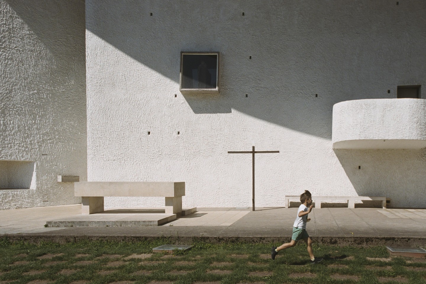 A boy runs across a grassy area in front of a white building with a cross and benches.