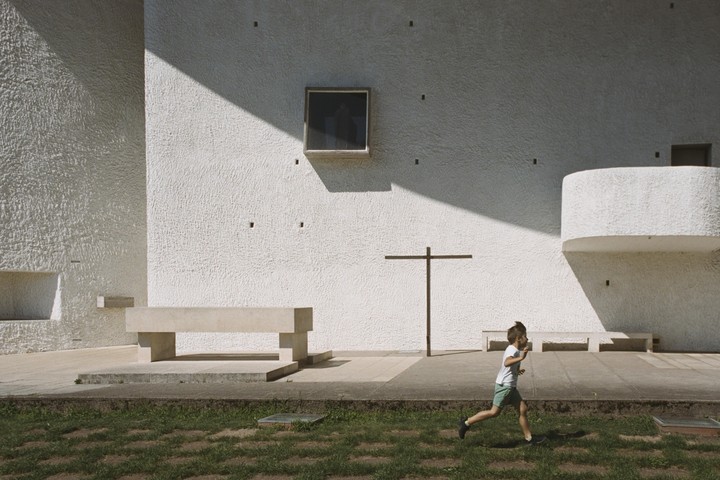 A boy runs across a grassy area in front of a white building with a cross and benches.