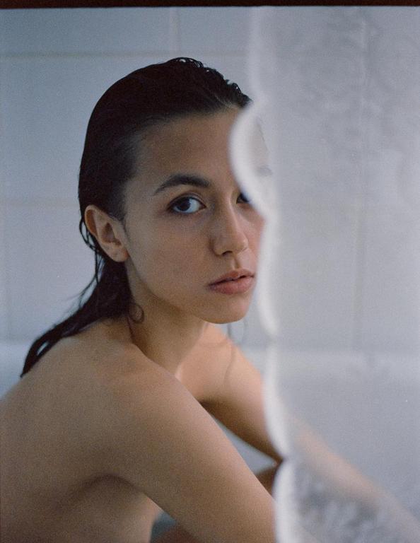 A woman with wet hair looks directly at the viewer from behind a shower curtain.