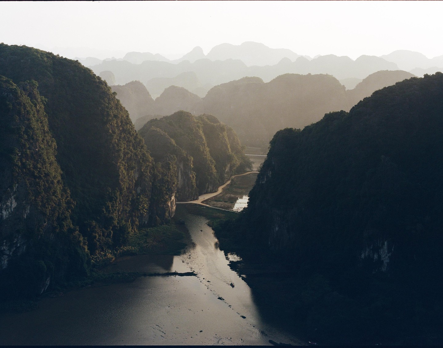A river flows through a valley surrounded by lush, steep mountains under a hazy sky.
