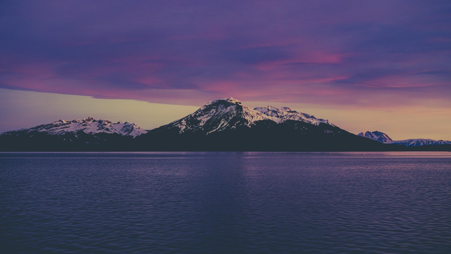 Snow-capped mountains rise above a body of water under a colorful sky at sunrise.