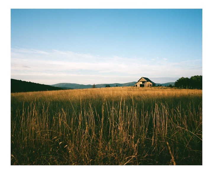 An old barn stands in a field of tall grass with mountains in the background under a clear sky.