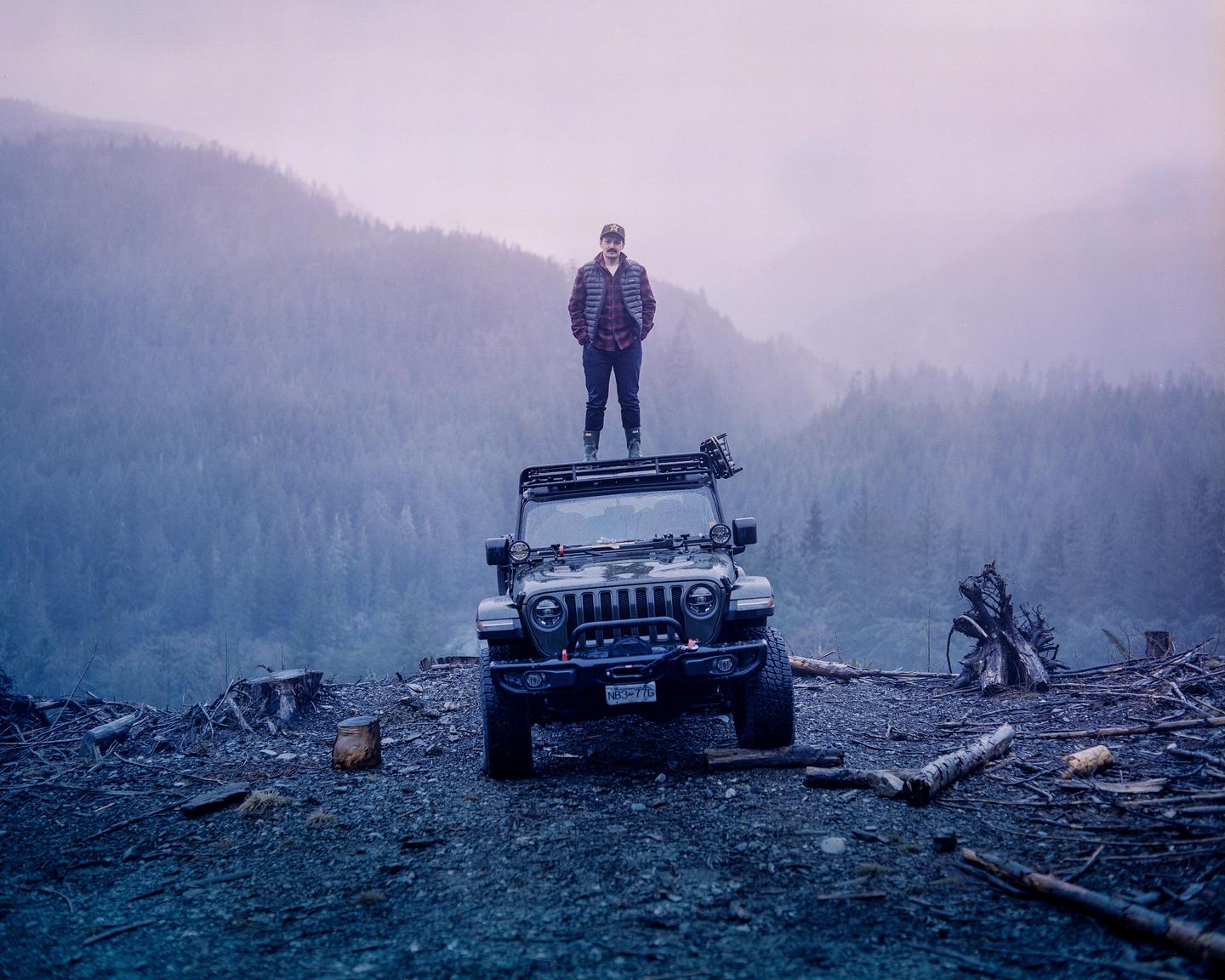 A man stands on top of a jeep in a deforested area with a forest and mountains in the background.