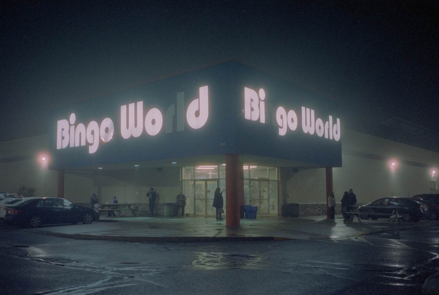 A building with a large "Bingo World" sign is illuminated at night, with several people standing outside and cars parked in the foreground.