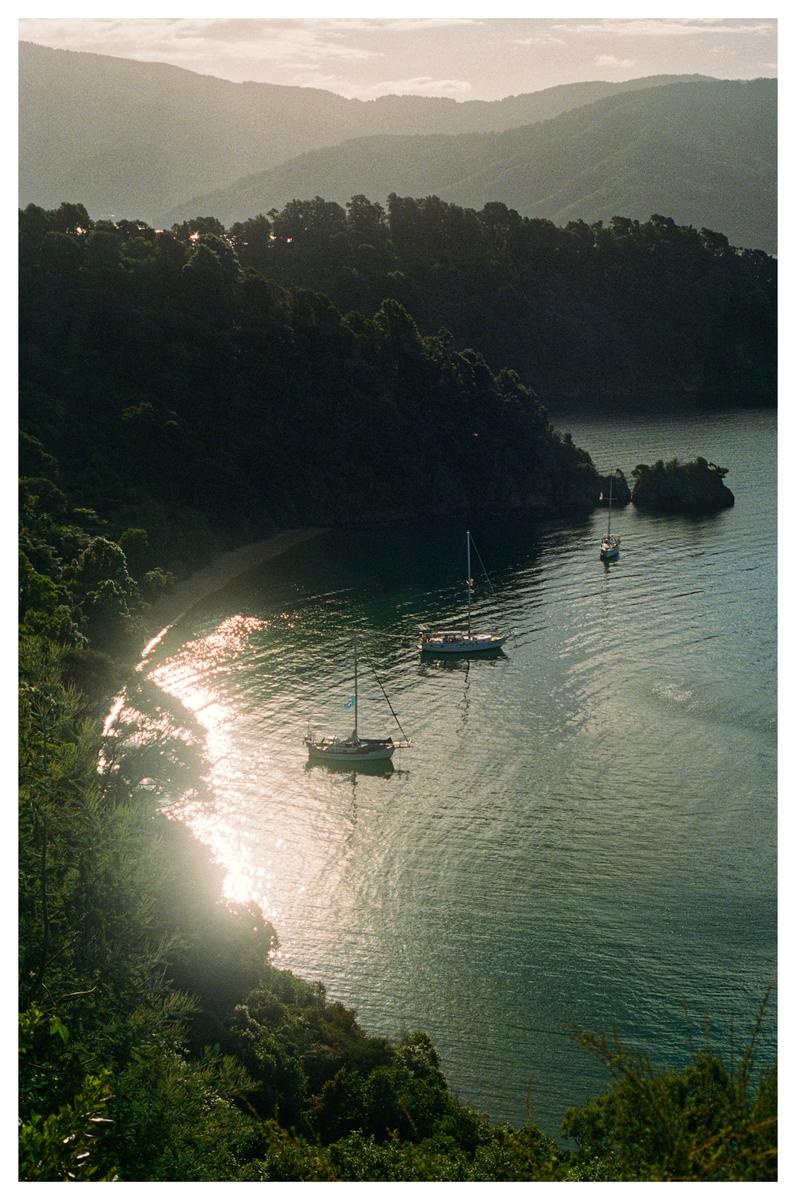 Three sailboats are anchored in a calm inlet surrounded by lush, forested hills under a hazy sky.
