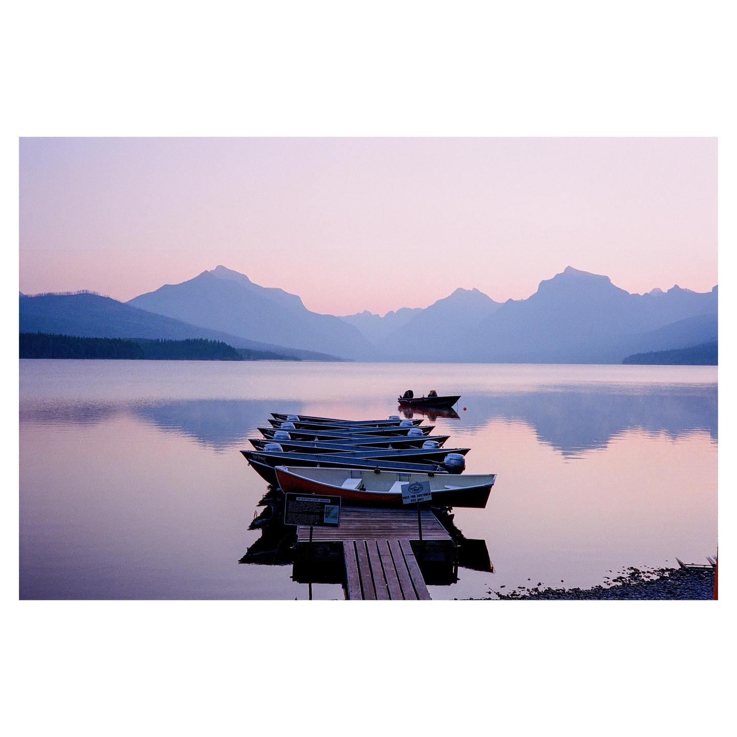 A line of boats is tied to a wooden dock on a lake with mountains in the background at dusk.