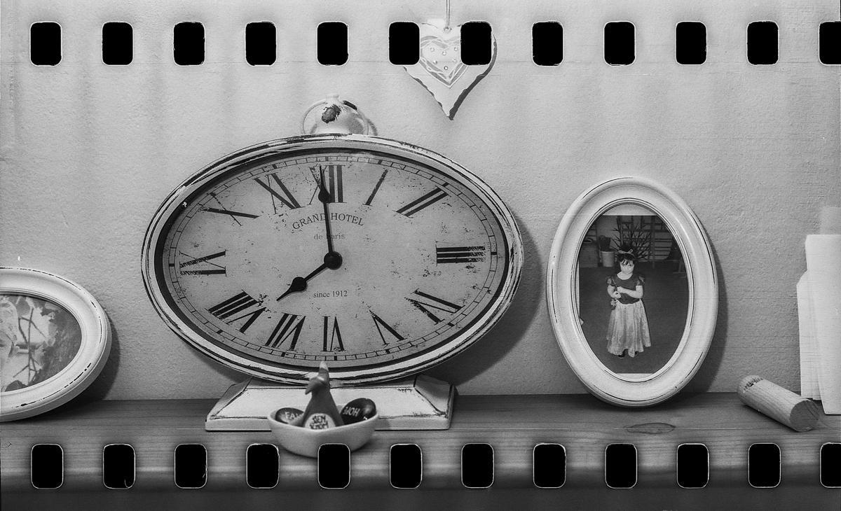 A large oval clock with Roman numerals sits on a wooden shelf next to a framed photo of a girl.