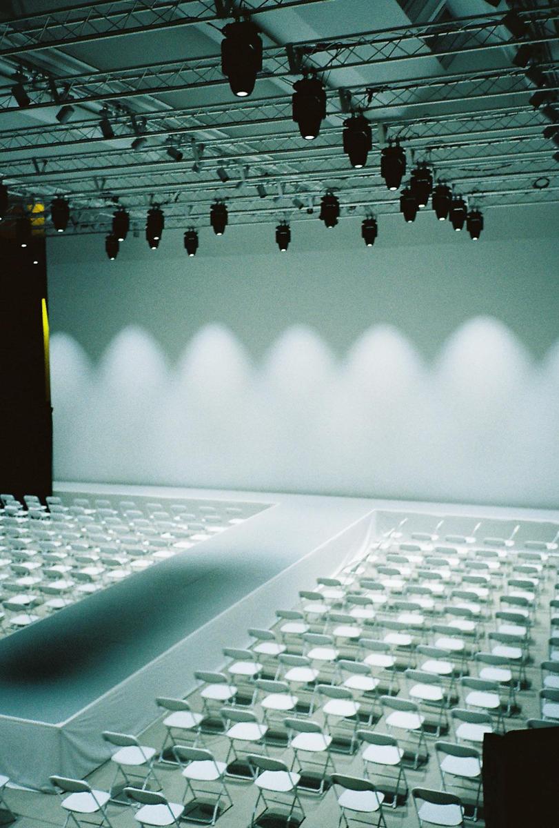 A fashion show runway is set up with rows of empty white chairs and spotlights illuminating a white wall.