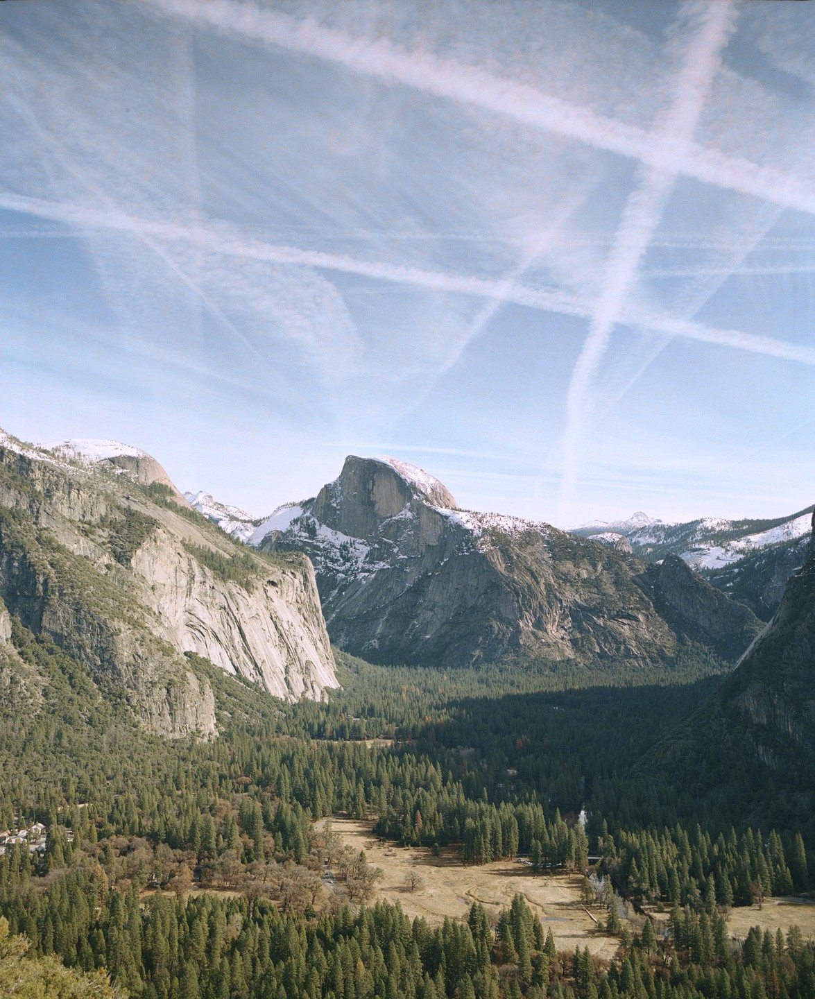 A wide view of Yosemite Valley shows Half Dome, snow-capped mountains, a forest, and contrails crisscrossing the sky.