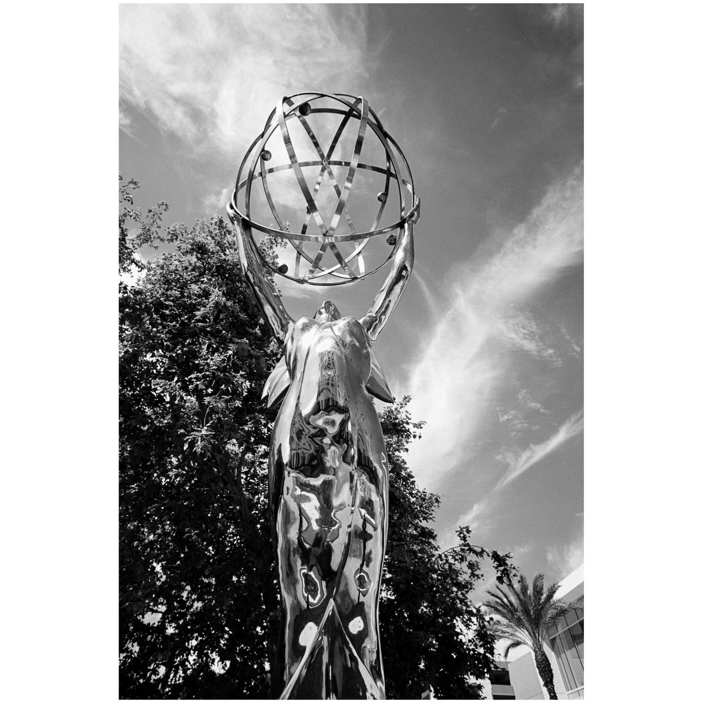 A low-angle black and white shot shows a statue of a woman holding a globe above her head, with trees and a cloudy sky in the background.