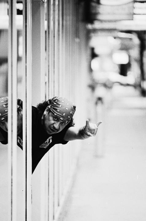 A man wearing a bandana and a t-shirt peeks from behind a white railing and makes a hand gesture.