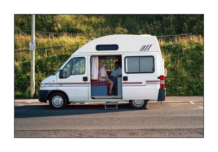 A white camper van with its side door open is parked on the side of a road, revealing a man and a woman sitting inside.