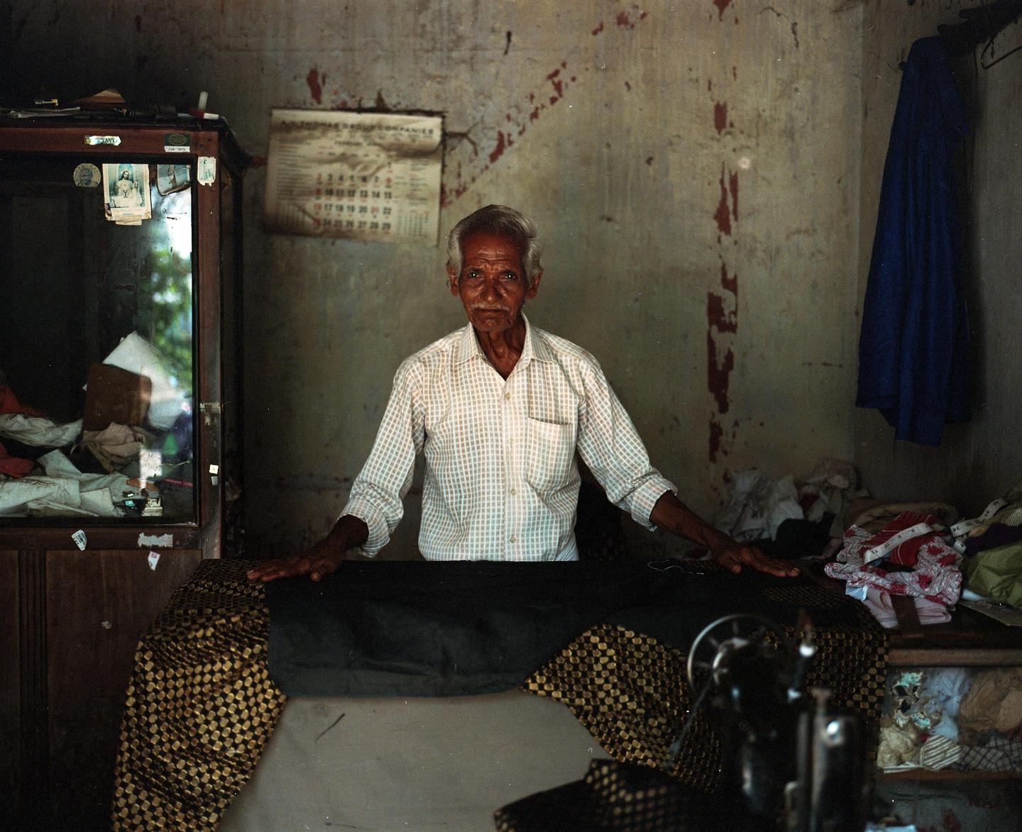 An elderly man with a mustache and white hair stands behind a table with fabric in a room with peeling paint.