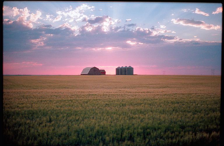 A barn and grain silos stand in a field under a pink and purple sunset.
