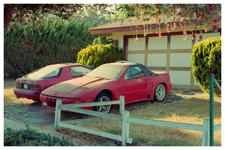 Two red sports cars are parked in a driveway next to a garage, with a white fence in the foreground.